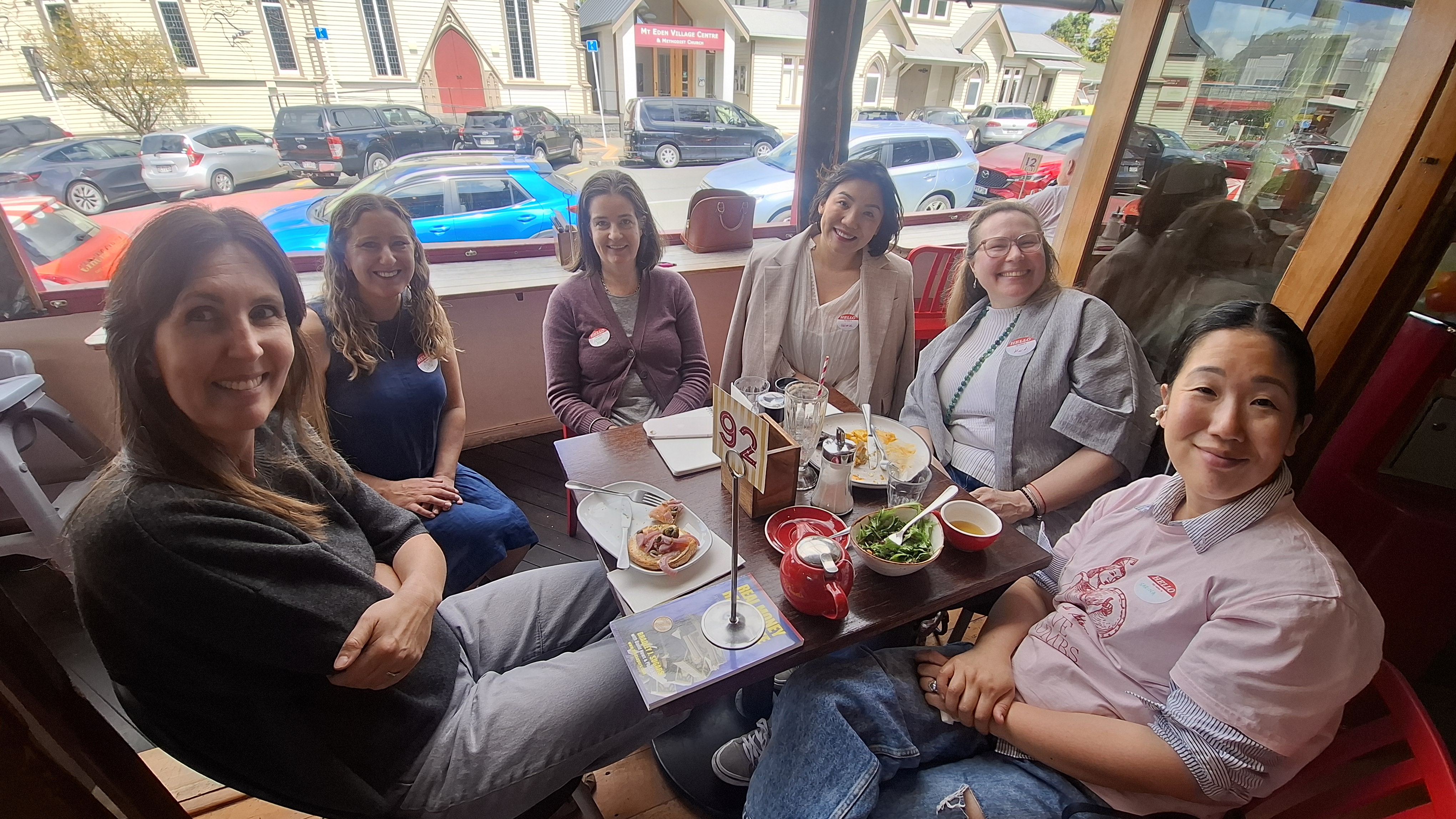 Six women smiling around a lunch table by the window at Circus Circus, Mt Eden, wearing name tags